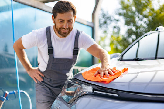 A smiling man in overalls is carefully drying the hood of a car with a bright orange microfiber cloth after a wash
