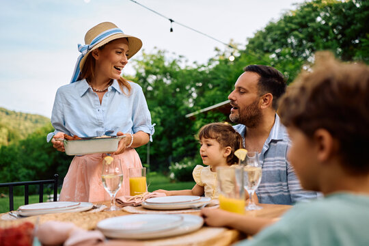 Happy woman brining food at table while having lunch with her family on patio.