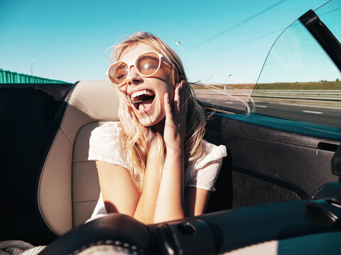 Portrait of young beautiful and smiling hipster female in convertible car. Sexy carefree woman driving cabriolet. Positive model riding and having fun in sunglasses at sunset
