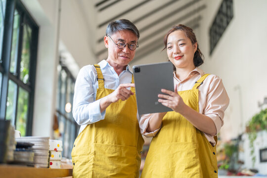 Senior happy asian man and young woman in yellow aprons using digital tablet in retail store, Family small business owners checking ceramic stock, Teamwork and SME boutique shop management concept