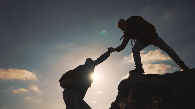 Hikers helping each other climb mountain peak at sunset. Teamwork silhouette shows strong support during outdoor climbing adventure. Friends assist partner reaching summit on rocky terrain with trust.