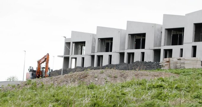 A row of modern, unfinished concrete houses on a construction site with an excavator, piles of gravel, and a grassy hill in the foreground.
