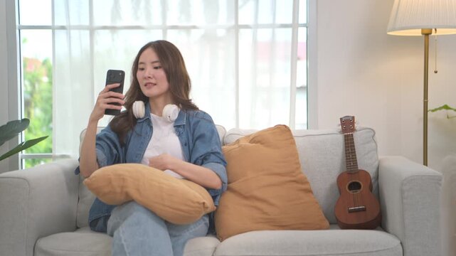 Young Asian woman relaxing on a sofa with a smartphone and headphones beside a ukulele in a bright living room