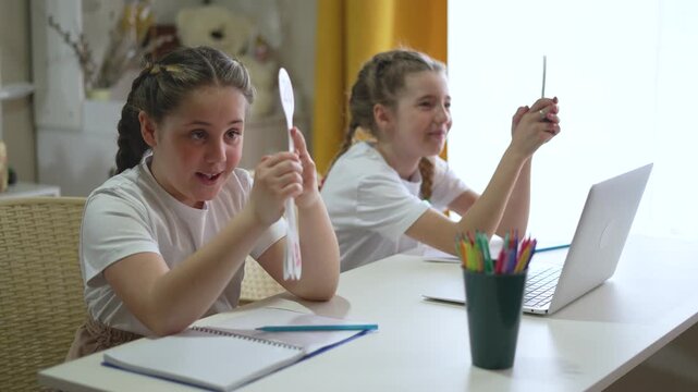 Two smiling girls learning online with laptop holding numbered cards at home. Happy students studying together. Sisters doing homework. Children education with computer. Kids learn at table.