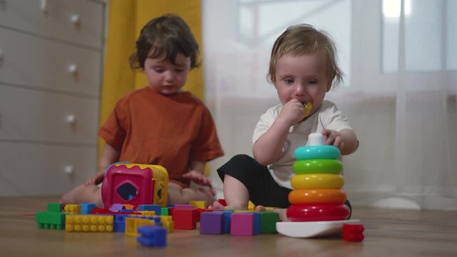Two toddlers play with colorful building blocks and stacking rings on bedroom floor. Children explore educational toys together. Kids share playtime with blocks and rainbow stacker toy.