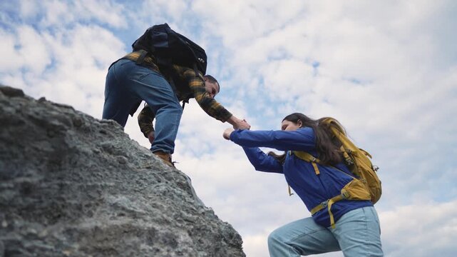 Hikers help each other climb rocky cliff with backpacks. Teamwork assists partner up mountain rock face. Friends support outdoor adventure climbing. Hiker pulls companion up steep terrain.