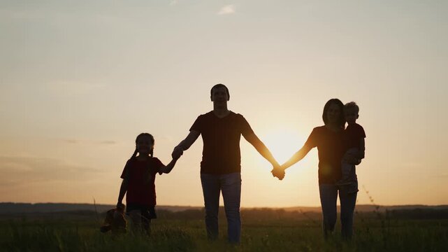 Happy family walking in field at sunset. Father and mother hold hands with children. Girl carries teddy bear while walking through grass. Family strolls together at golden hour in open meadow.