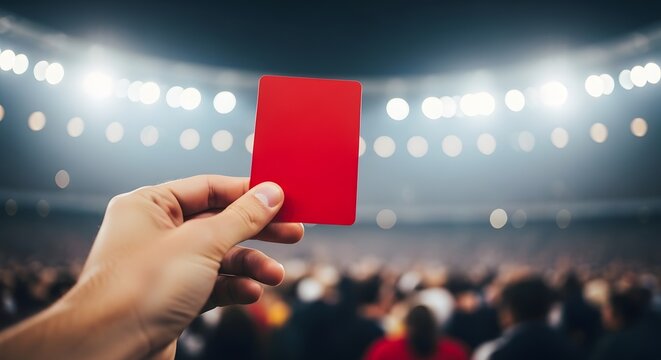 Red card being held up by hand in sports stadium with blurred crowd and bright lights in background