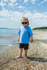 Child stands on beach near water with blue sky overhead in daytime while wearing sunglasses and a cap © DSMT