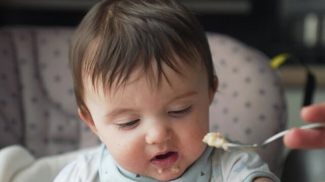 Baby eating messy food with spoon in highchair during feeding time. Parent feeds infant porridge. Toddler learns eating solid food. Child has messy meal with food on chin developing feeding skills.
