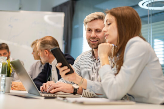Two business people discussing work together using a smartphone in a modern office, colleagues on the background
