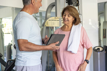 Obraz premium Mature man holding tablet instructing senior woman wearing pink tee with towel and watch at gym