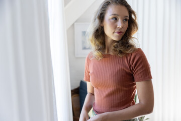 Naklejka premium Woman standing by bright window at home, gazing right, wearing ribbed rust top, curtains visible
