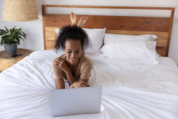 Obraz premium African American woman lying on white bed smiling while using laptop with potted plant in bedroom