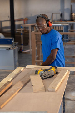 Woodworker is holding nail gun and aligning wooden plank at workbench in bright carpentry workshop