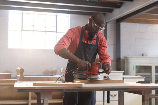 Carpenter wearing safety goggles sanding wood plank on workbench in workshop near window light