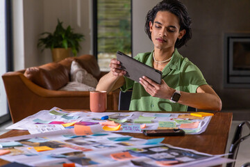Obraz premium Asian man sitting at wooden table in living room holding tablet, reviewing sticky notes, photos