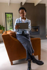 Obraz premium African American woman in denim jacket sitting on arm of leather armchair holding tablet at home
