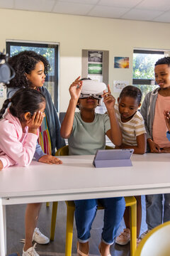 Diverse children gathering around classroom table seated child adjusting white VR headset, tablet