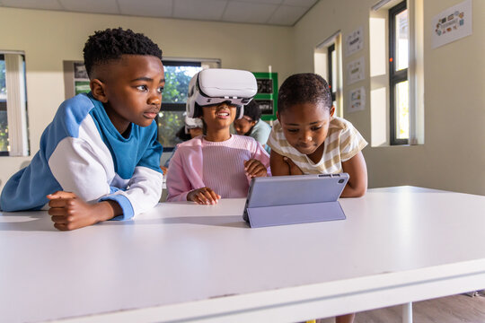African American children trying virtual reality and using tablet at classroom table with headset