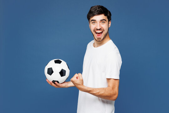 Young man fan wears basic white t-shirt cheer up support football sport team hold in hand soccer ball watch tv live stream doing winner gesture celebrate clench fist isolated on plain blue background.