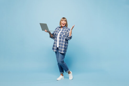 Full body elderly fun blonde IT woman 50s years old wear checkered shirt casual clothes hold use work on laptop pc computer look aside isolated on plain pastel light blue background Lifestyle concept