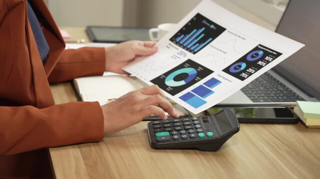 Close view of a business person reviewing printed charts while using a calculator at a desk with a laptop, showing budgeting, finance, analysis and office work.