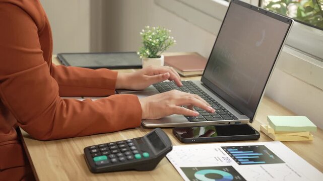 Woman in a brown blazer typing on a laptop at a wooden desk near a window, with calculator, phone, sticky notes, tablet and financial charts in a bright home office