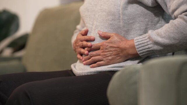 Senior woman sitting on a sofa and holding her stomach in pain at home, showing abdominal discomfort, indigestion, or illness in a calm indoor setting.