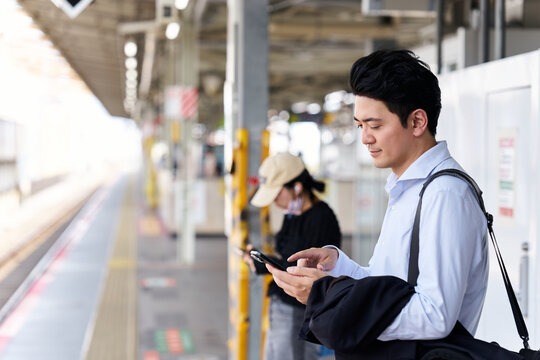 駅のホームでスマートフォンを操作する30代の日本人男性会社員と背景にいる女性
