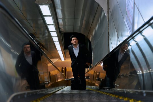 スーツを着てキャリーケースを持つ30代の日本人男性会社員が駅のエスカレーターに乗る様子