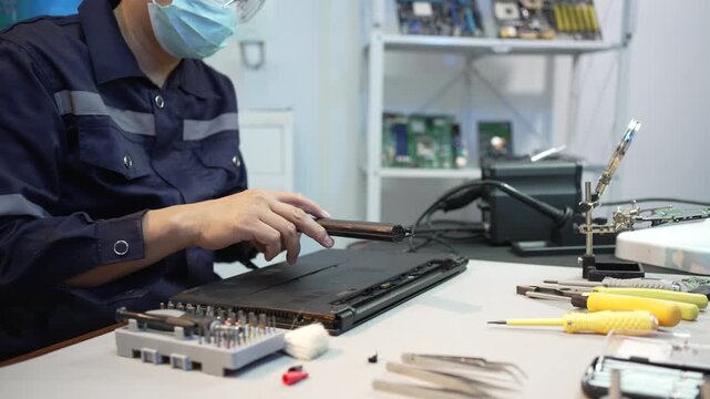 Technician repairs a laptop at a workbench using a screwdriver, with tools and electronic parts spread across the table in a clean workshop