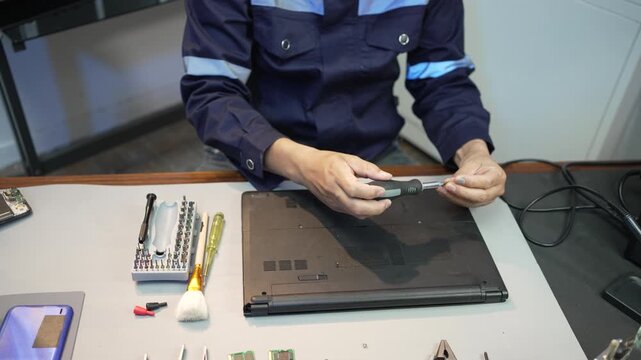 Technician repairs a laptop at a workbench using a screwdriver, with tools and electronic parts spread across the table in a clean workshop