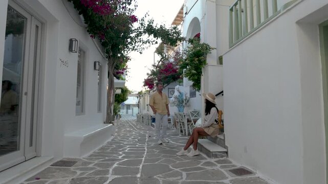A couple wanders through the picturesque streets of Paros, Greece, where vibrant flowers bloom and traditional architecture enchants visitors. Experience the beauty of this Cycladic island