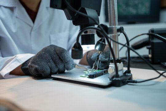 Technician in a lab coat and mask repairs a small circuit board under a digital microscope at a workbench, showing electronics testing, research, and precision work.