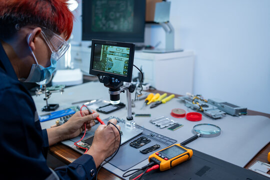 Technician repairing a smartphone at a workbench using a microscope screen and small tools in an electronics lab.