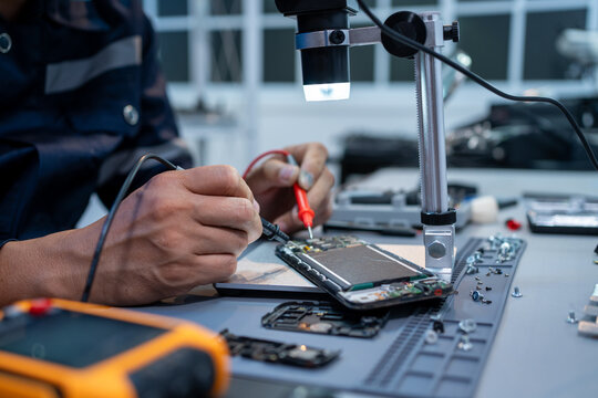 Technician repairing a smartphone at a workbench using a microscope screen and small tools in an electronics lab.