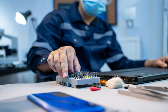 Masked repair technician works at a desk with small tools, phone parts and a laptop in a clean electronics service room.