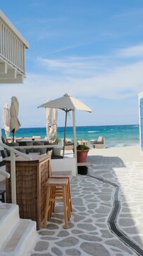 Experience the serene beauty of a beachside cafe in Paros, Greece. Soft waves lap against the shore as sunlight bathes the area in warmth. Enjoy the perfect holiday vibe.