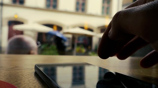 Close up view of a hand scrolling through a social media feed on a smartphone at a cafe table, with a blurred city street and people walking under umbrellas in the rainy background