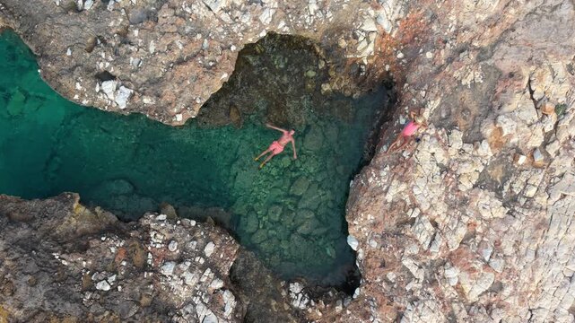 Discover the stunning coastal beauty of Paros, Greece, where vibrant waters meet rocky shores. A relaxes by the ocean natural pool, a couple of man and a woman from above drone view