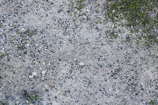A rustic dirt path or walkway. The coarse, grainy texture consists of fine light grey sand heavily scattered with small, multi-colored pebbles, gravel chips, and crushed stones.