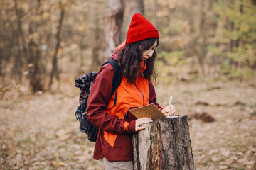 Fototapeta premium Woman stands in autumn forest while writing in notebook on tree stump. Biological resource monitoring, sustainable forest management, ecological auditing process.