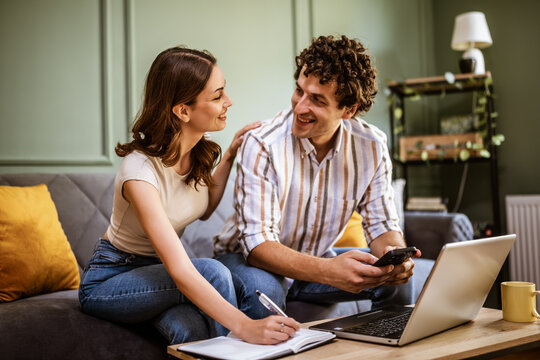 Adult couple is sitting at sofa at home and using laptop. They are shopping online and making shopping list.