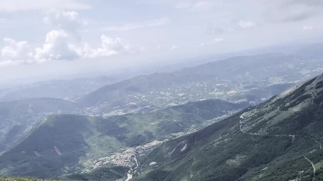 Vista dalla cresta di una montagna, con la camera che mostra pendii verdi, strade serpeggianti e valli profonde che si estendono verso rilievi lontani sotto un cielo parzialmente nuvoloso