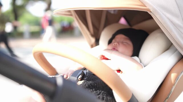 Soft sunlight bathes a sleeping infant. Baby rests peacefully in a stroller's cushioned seat. Tiny hands curl near the face in quiet slumber. Wooden handle grips the stroller's frame