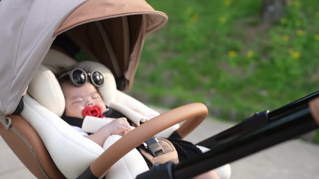 Cute baby rests peacefully in stroller. Baby wears sunglasses and sips red pacifier. Soft tones and gentle motion create calm atmosphere. Perfect for parenting, lifestyle, or baby product content
