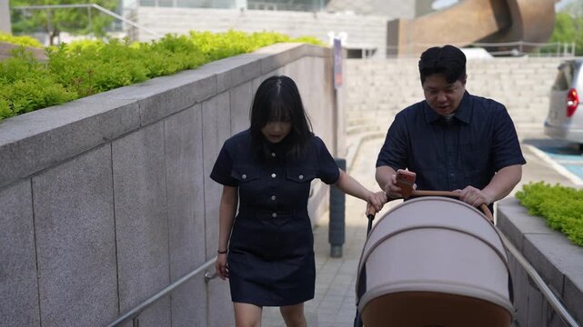 Couple walks down urban pathway with baby stroller. Woman looks down, man smiles gently while pushing stroller. Natural light shows calm, everyday parenting moment. Ideal for parenting, family