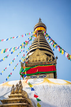 A stunning low-angle photograph of the sacred Boudhanath Stupa in Kathmandu, Nepal, featuring the iconic Buddha Eyes, colorful Tibetan prayer flags, a gilded spire, and the massive white dome beneath 