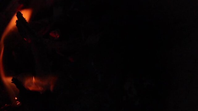 Close-up of small orange campfire flames and glowing embers in darkness, creating a warm, atmospheric fire background with dramatic lighting and copy space.